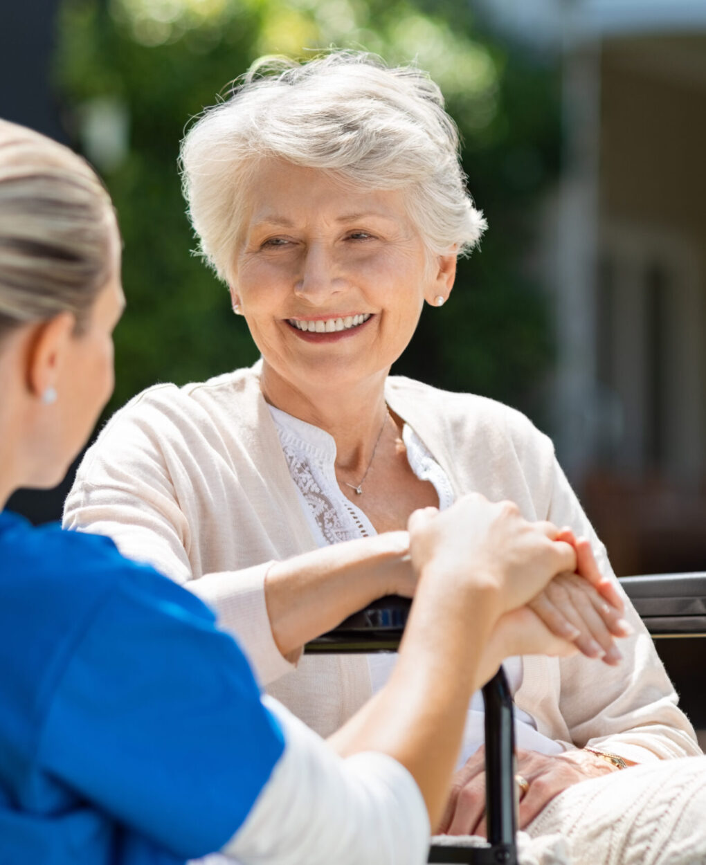 Smiling senior patient sitting on wheelchair with nurse supporting her. Doctor looking at elderly patient on a wheelchair in the garden. Nurse holding hand of mature woman outside pension home.